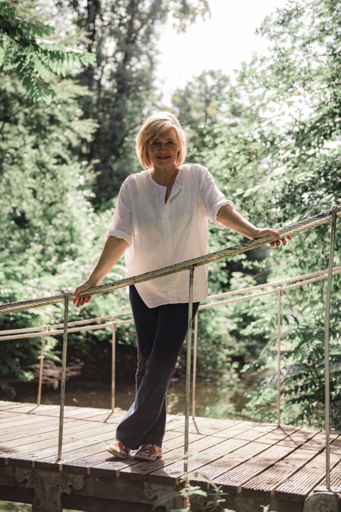 Een persoon in een wit shirt staat op een brug in een weelderig groen bos, leunend tegen de reling en glimlachend, wat de positiviteit van de natuur belichaamt.