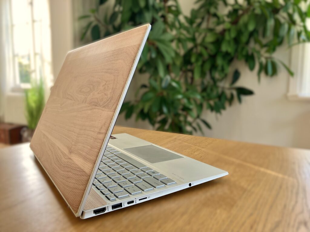 Een laptop met een houten afwerking op het deksel staat opengeslagen op een houten tafel en vormt een harmonieus geheel met de wazige groene plantenachtergrond. Echt een positief iets.