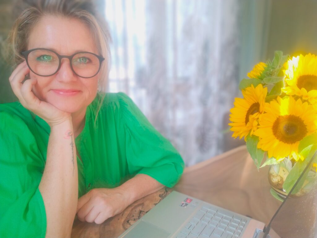 Een persoon in een groen shirt met een bril glimlacht en straalt warmte uit, terwijl zij aan een tafel zit met een laptop en een vaas met zonnebloemen.